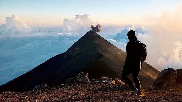 The YouTube thumbnail of my 2021 film Hiking to Acatenango Volcano in Guatemala: me on the rim, back to camera, Fuego's cone puffing ash behind me over a sea of clouds